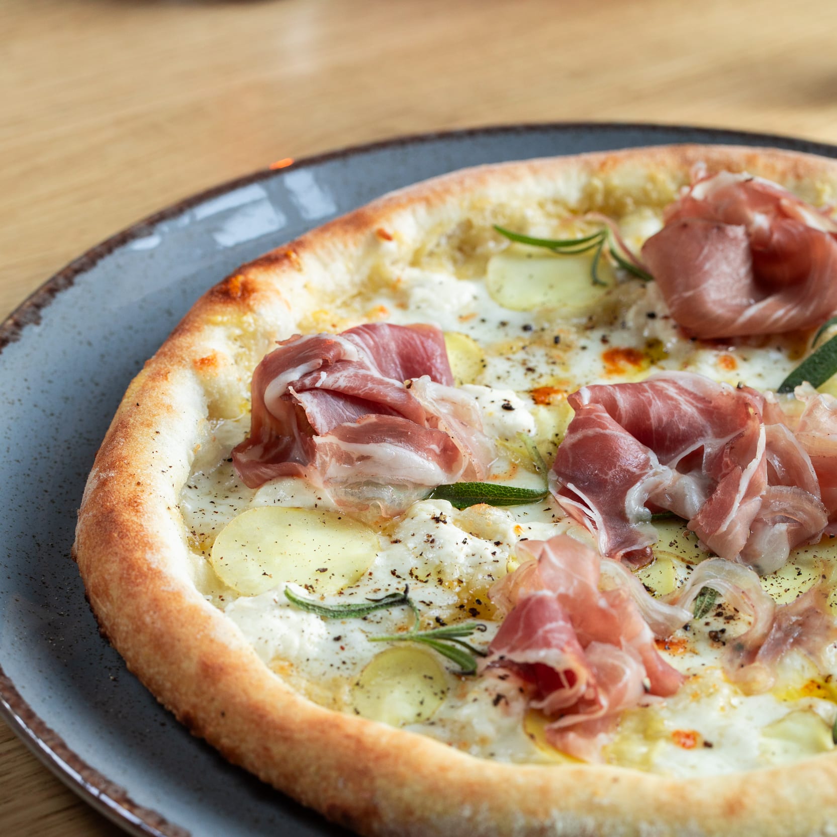 A close-up of a pizza on a plate, featuring ham, potatoes, and rosemary, with a golden crust, on a wooden surface.
