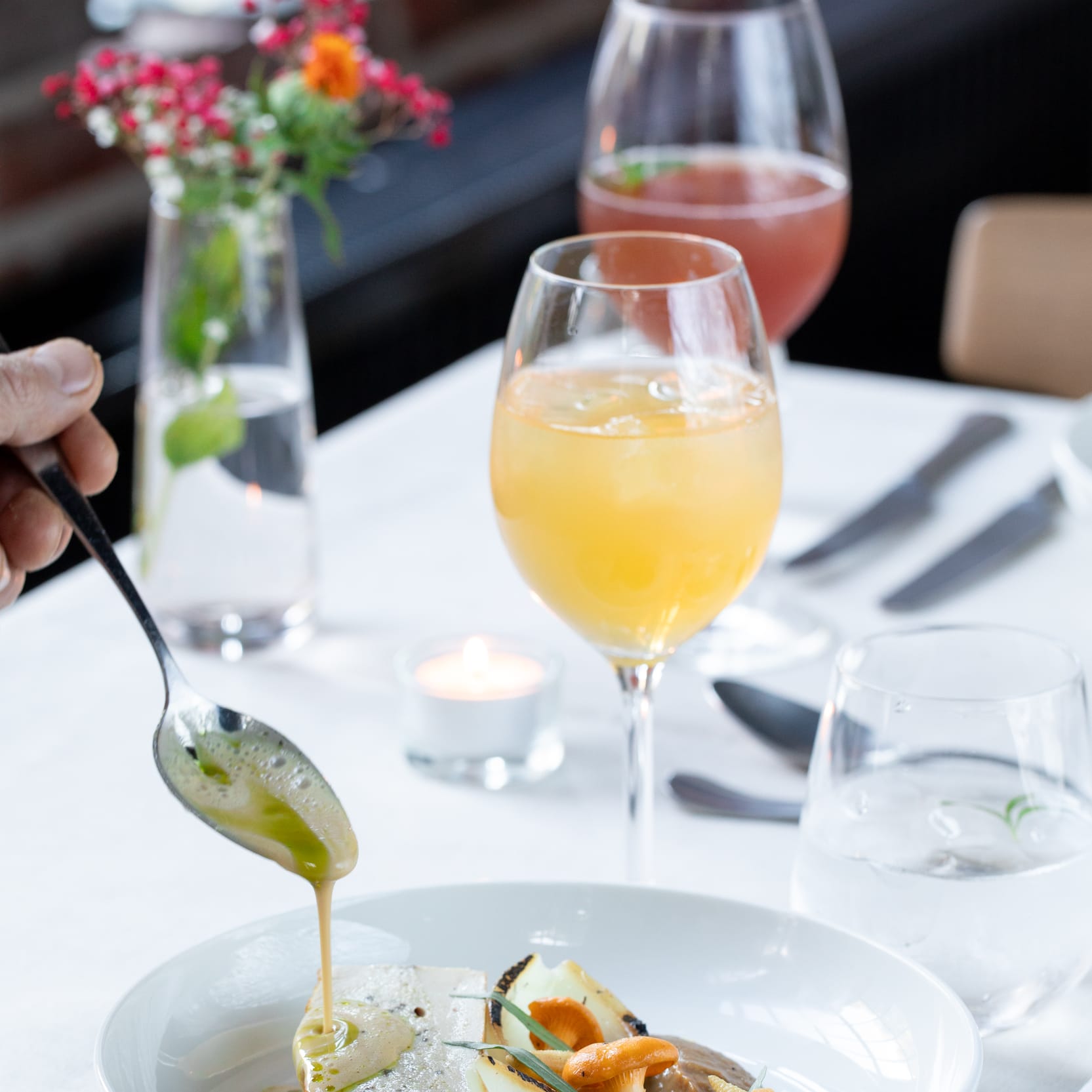 Close-up of a chef pouring sauce onto a plate of food with fish, mushrooms, and other vegetables in a restaurant setting. Drinks visible in background.