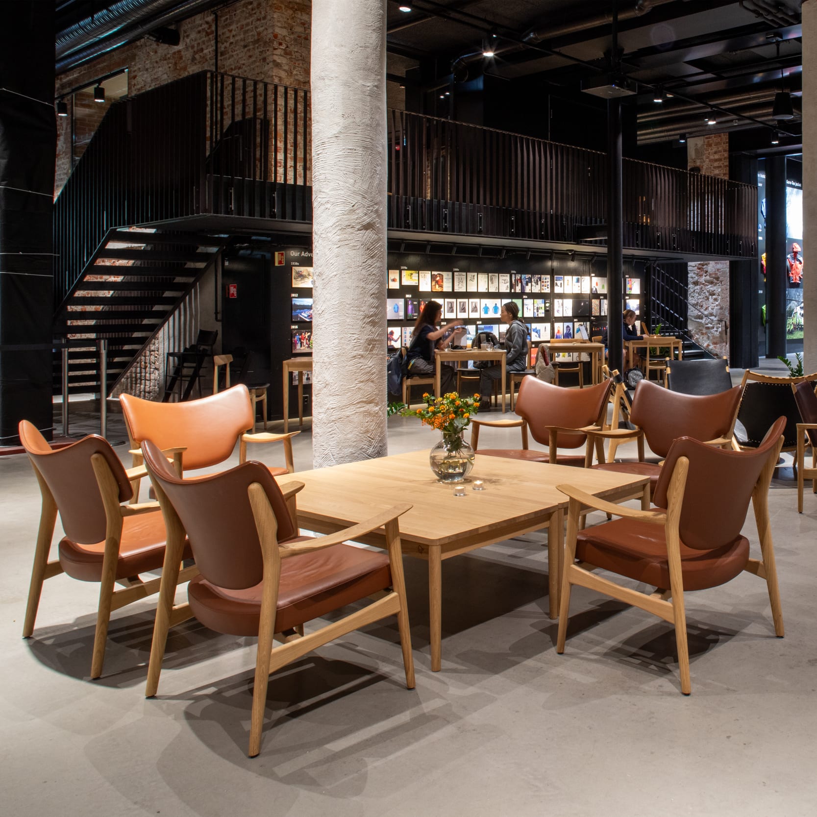 An open-concept interior with tables and leather chairs. People are seated at some tables. The design features exposed brick, pipes, and pillars.