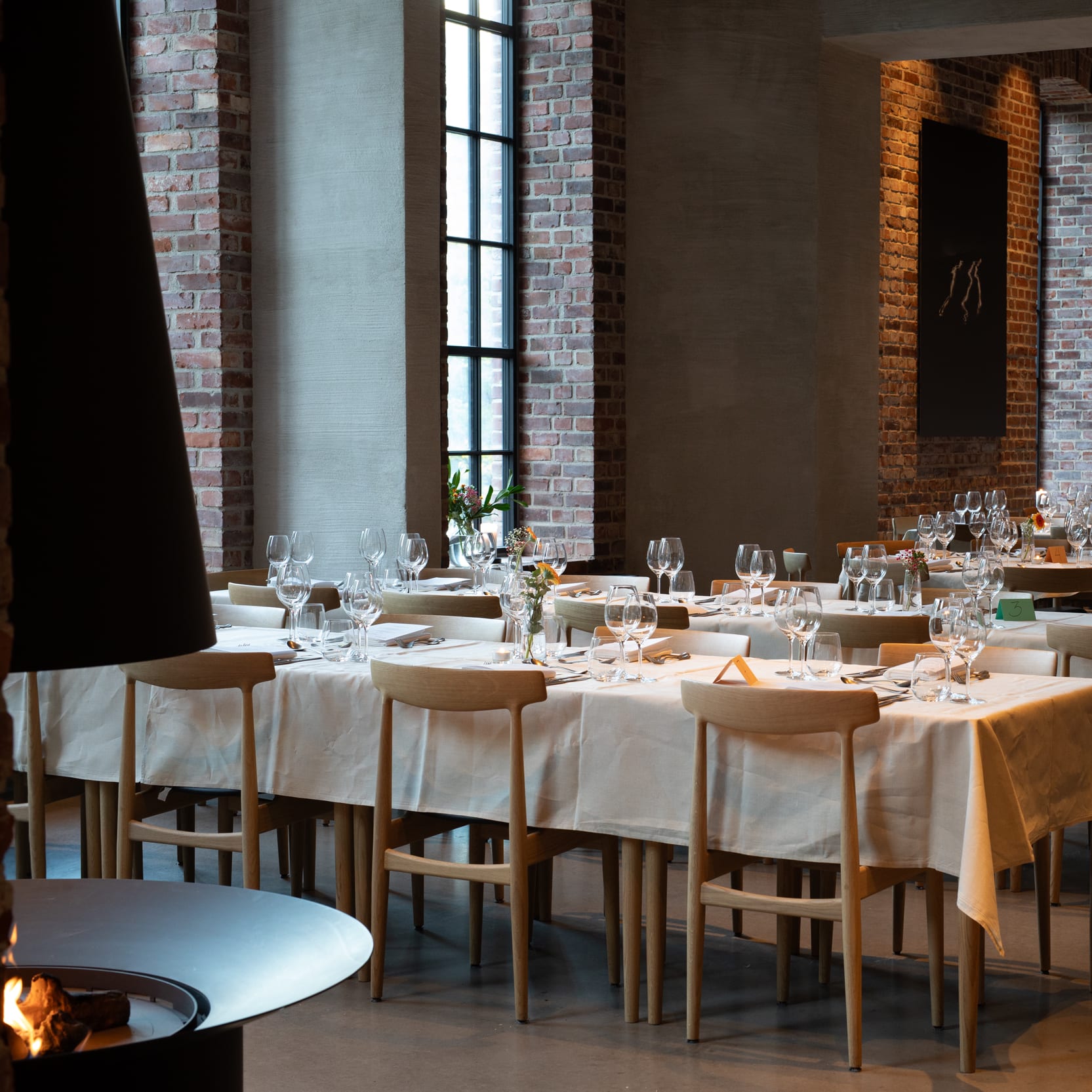 A wide shot of a dining room, set with tables for a formal event. The room features brick walls and large windows.