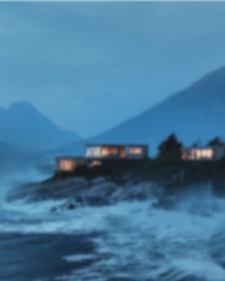 Illuminated modern cabins on a rocky coast with crashing waves and mountains at twilight.
