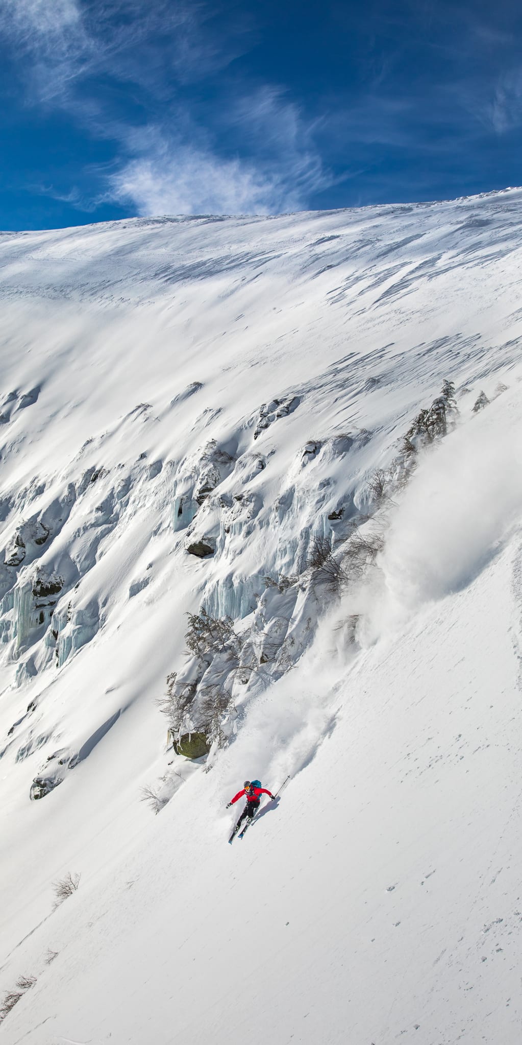 A skier in red Norrøna gear makes a turn down a snowy mountain slope. The skier is leaving a trail of snow behind them.