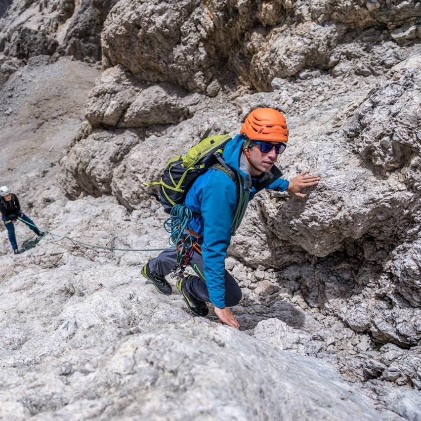 Two climbers on a steep, rocky mountain face. The man in foreground wears Norrøna jacket, orange helmet, backpack and rope, while another person climbs below.
