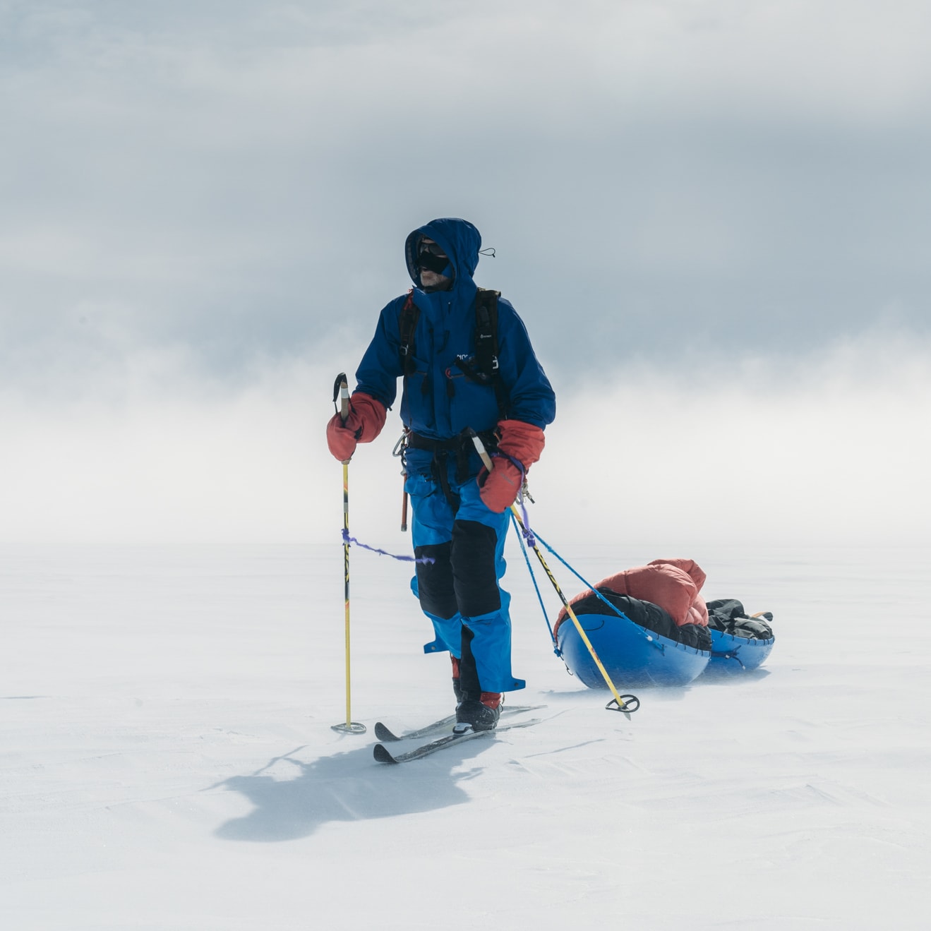 A person in Norrøna ski gear skis across a snowy landscape pulling a blue sled with gear. Overcast skies.
