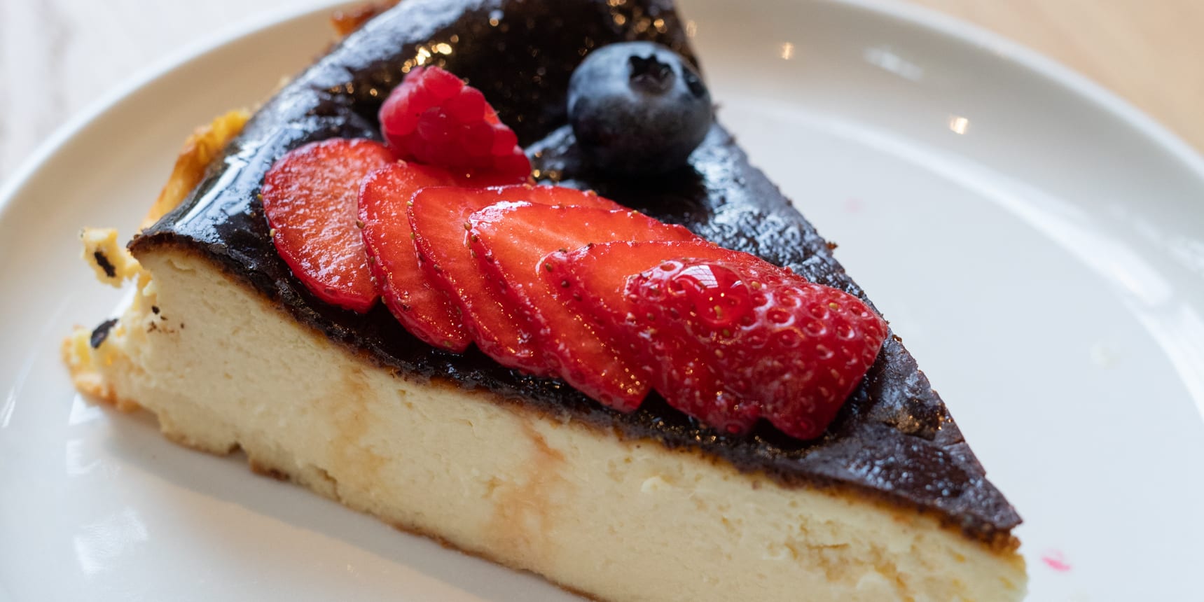 A slice of burnt Basque cheesecake with sliced strawberries, a raspberry, and a blueberry on a white plate.