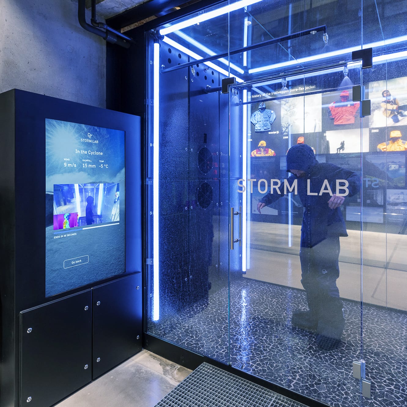 A person in a Norrøna jacket stands inside a glass enclosure, looking out at a display showing weather conditions and the history of Norrøna jackets.