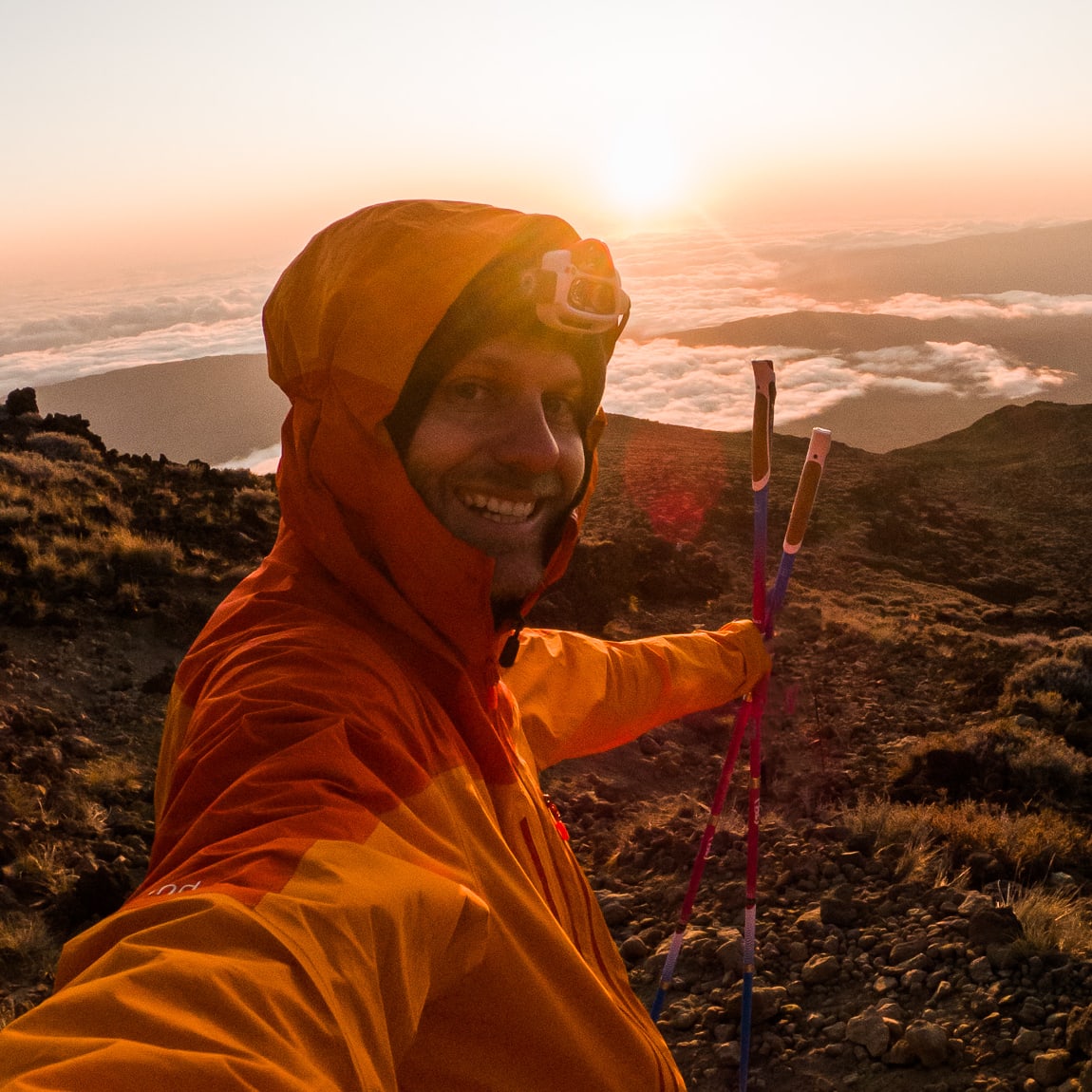 A person wearing a Norrøna orange jacket, smiling, holding trekking poles, stands on a mountaintop with a beautiful sunrise in the background.