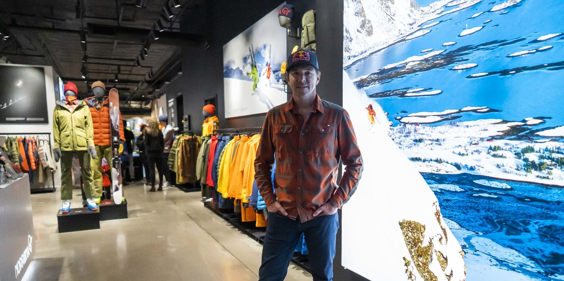 A man in a red and black shirt stands in a Norrøna store. Behind him is a large photo of a snowy mountain range and a body of water. 
