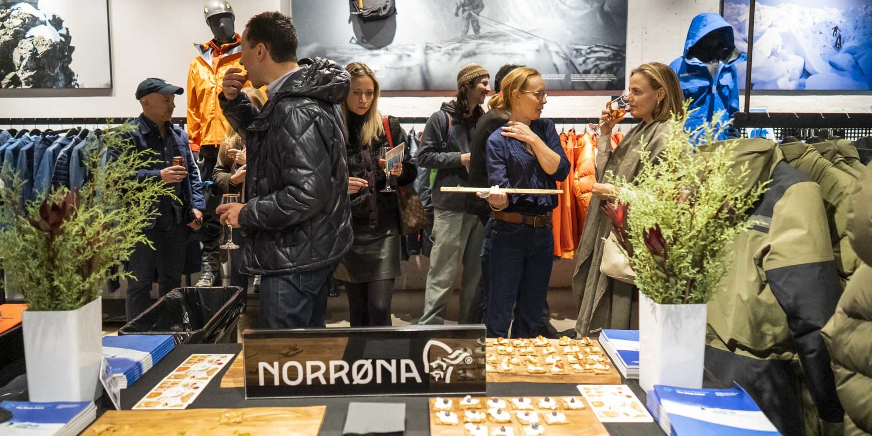 People gather in a Norrøna store, enjoying refreshments near a display of clothing and gear. 
