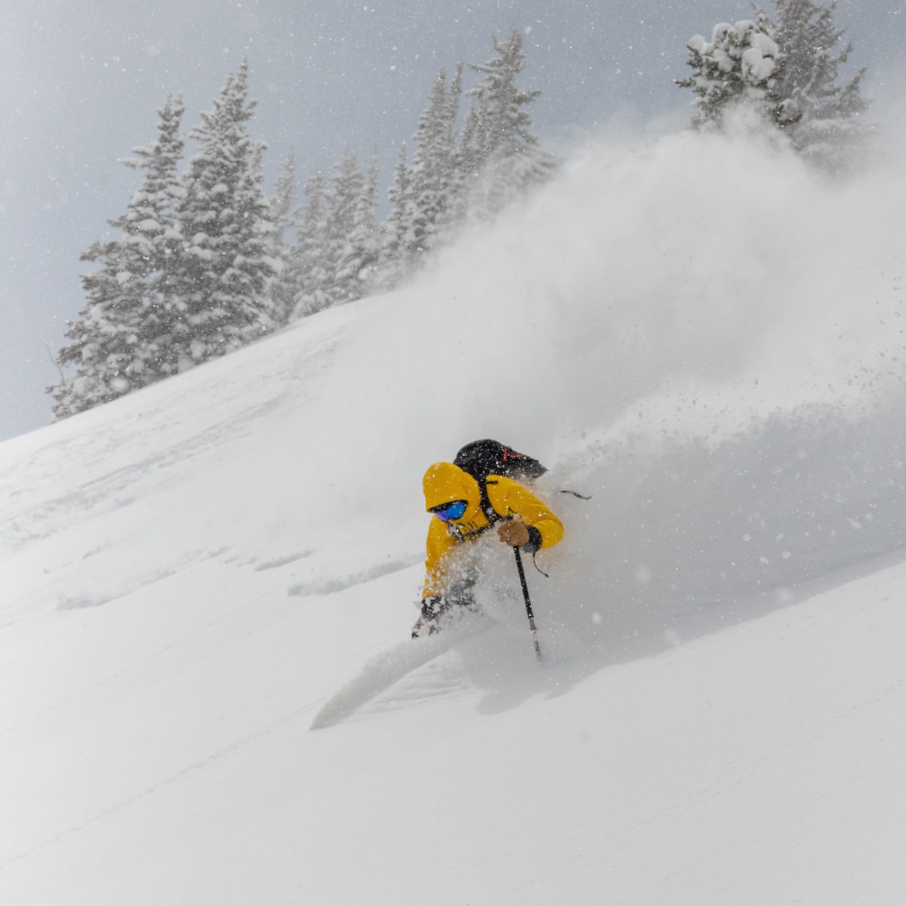 A skier in a Norrøna yellow jacket skis down a snowy slope, creating a spray of snow against a backdrop of snow-covered trees in a snowstorm.