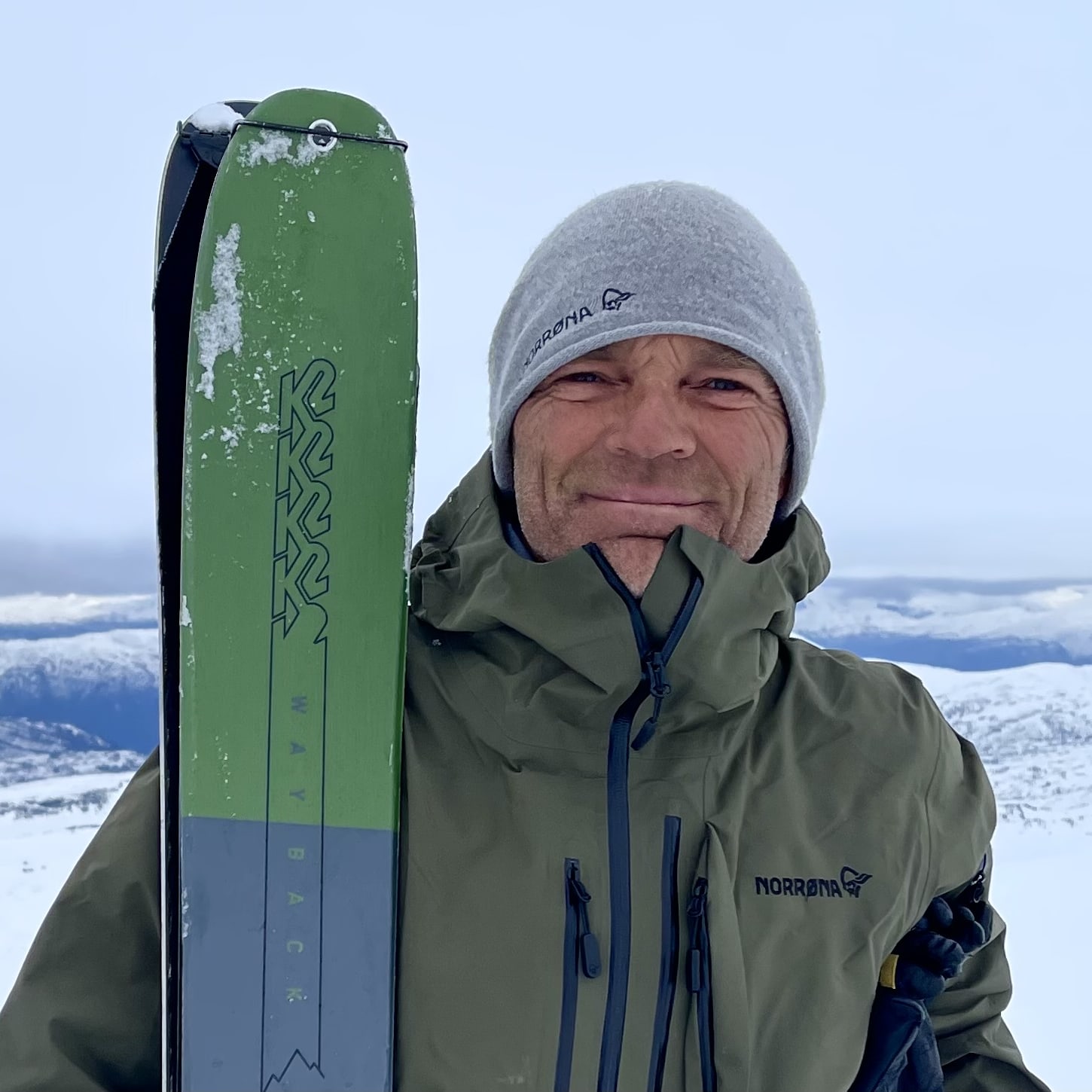 A man in a Norrøna jacket and hat holds a ski in a snowy mountain landscape. 
