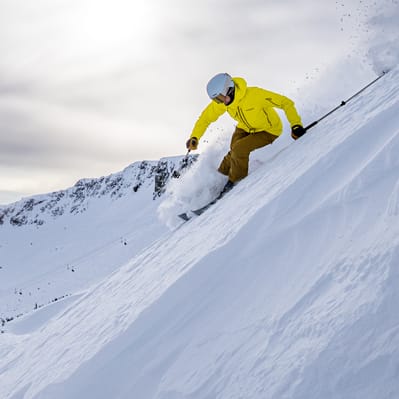A skier in a yellow jacket, wearing a helmet, is skiing down a snowy mountain slope.