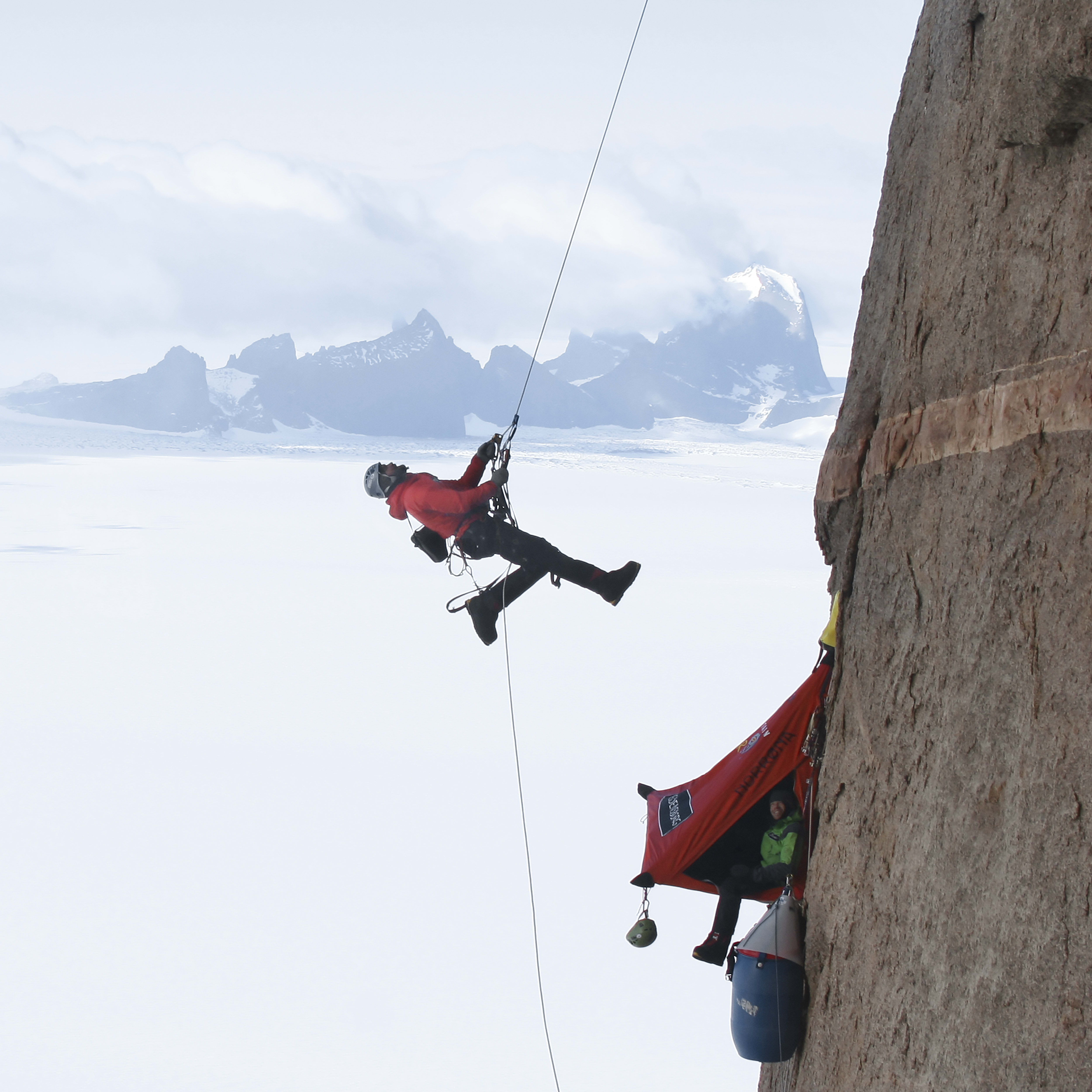 A person rappelling down a rock face in front of a snow-covered landscape.  The person is wearing a red jacket and black pants. A red tent is visible on the rock face.  A green climbing nut hangs below the tent.