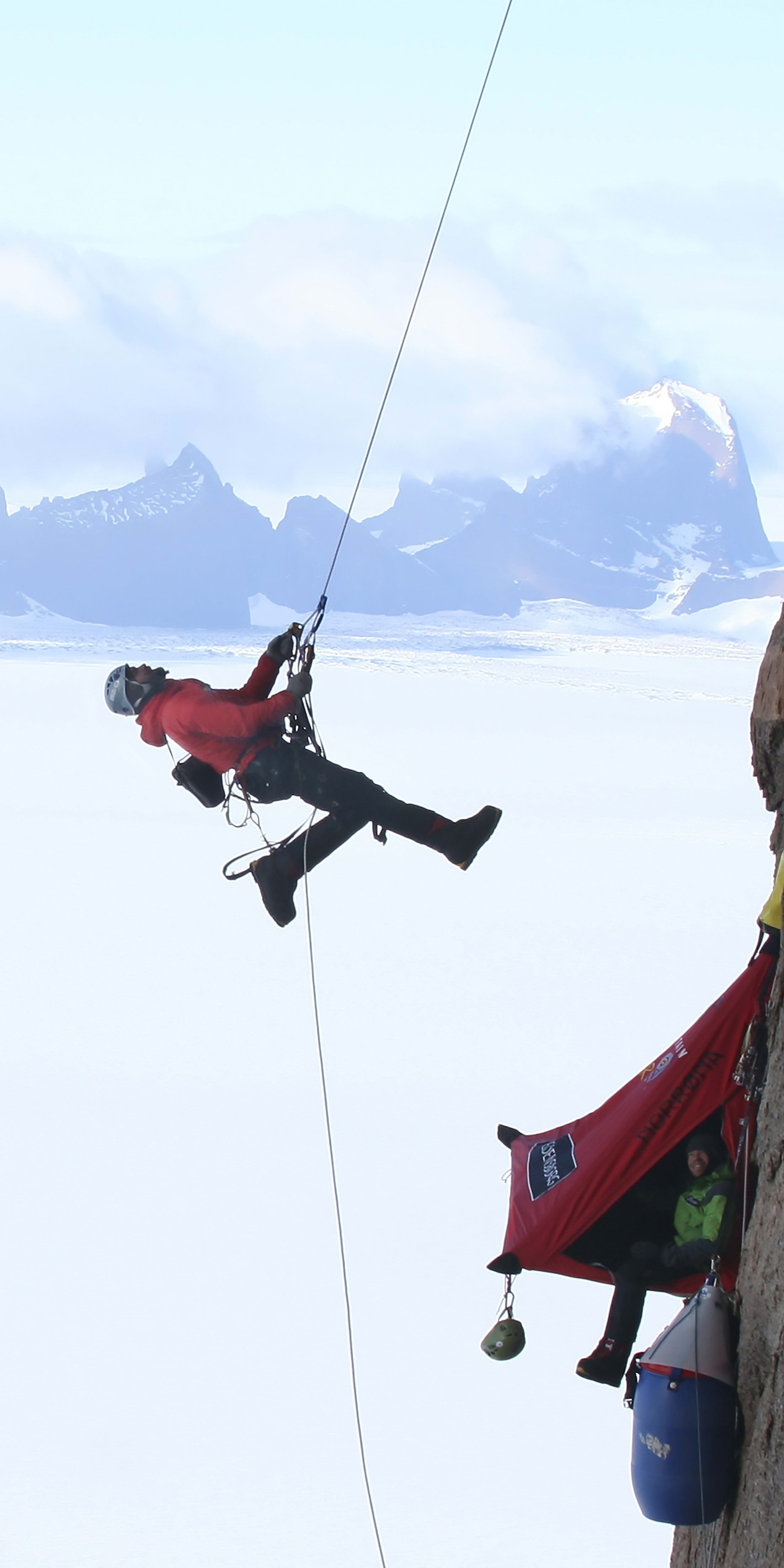A man in a red jacket and helmet is rappelling down a cliff face with a rope. He is in the middle of his descent, with a small, red tent hanging from the cliff face below him. The background is a snowy, mountainous landscape.