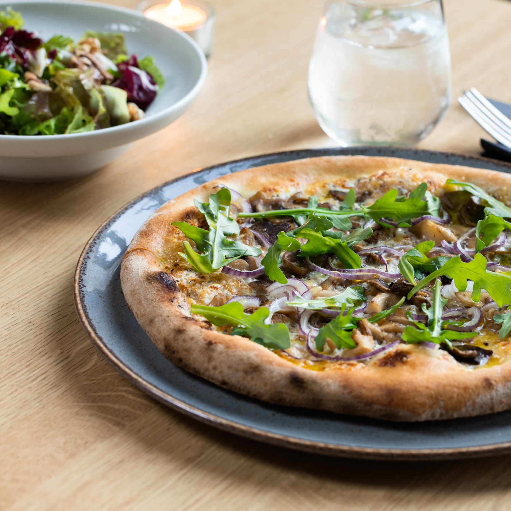 A pizza with arugula and red onion on a plate, next to a salad and glass of water.