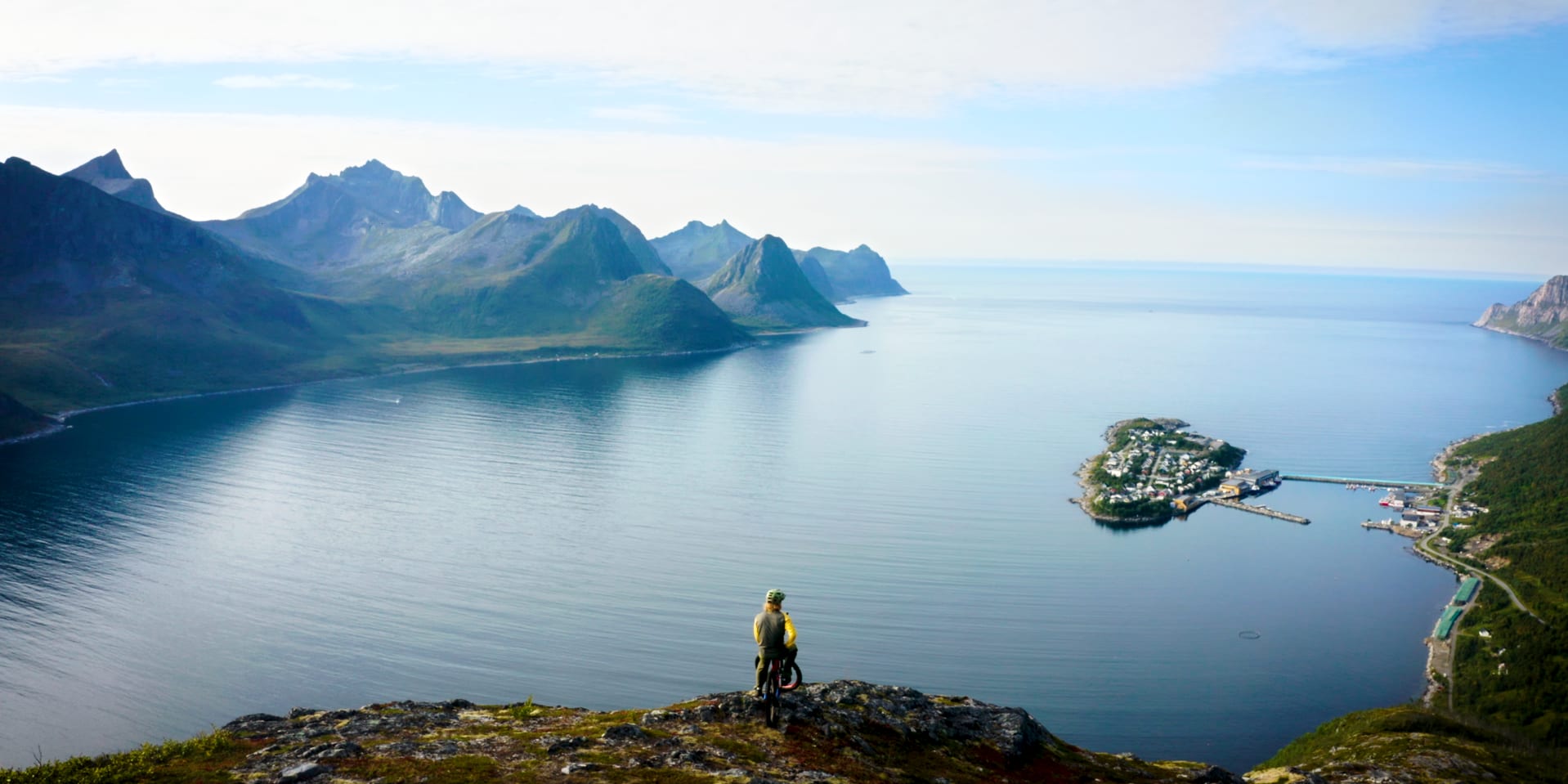 Cyclist in Norrøna gear looks out over a vast fjord, dramatic mountains, and a small island village.