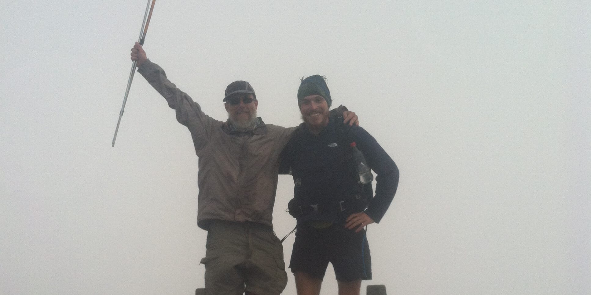 Two hikers stand on Katahdin, the highest peak in Maine, holding trekking poles. They are wearing Norrøna clothing and a backpack. 
