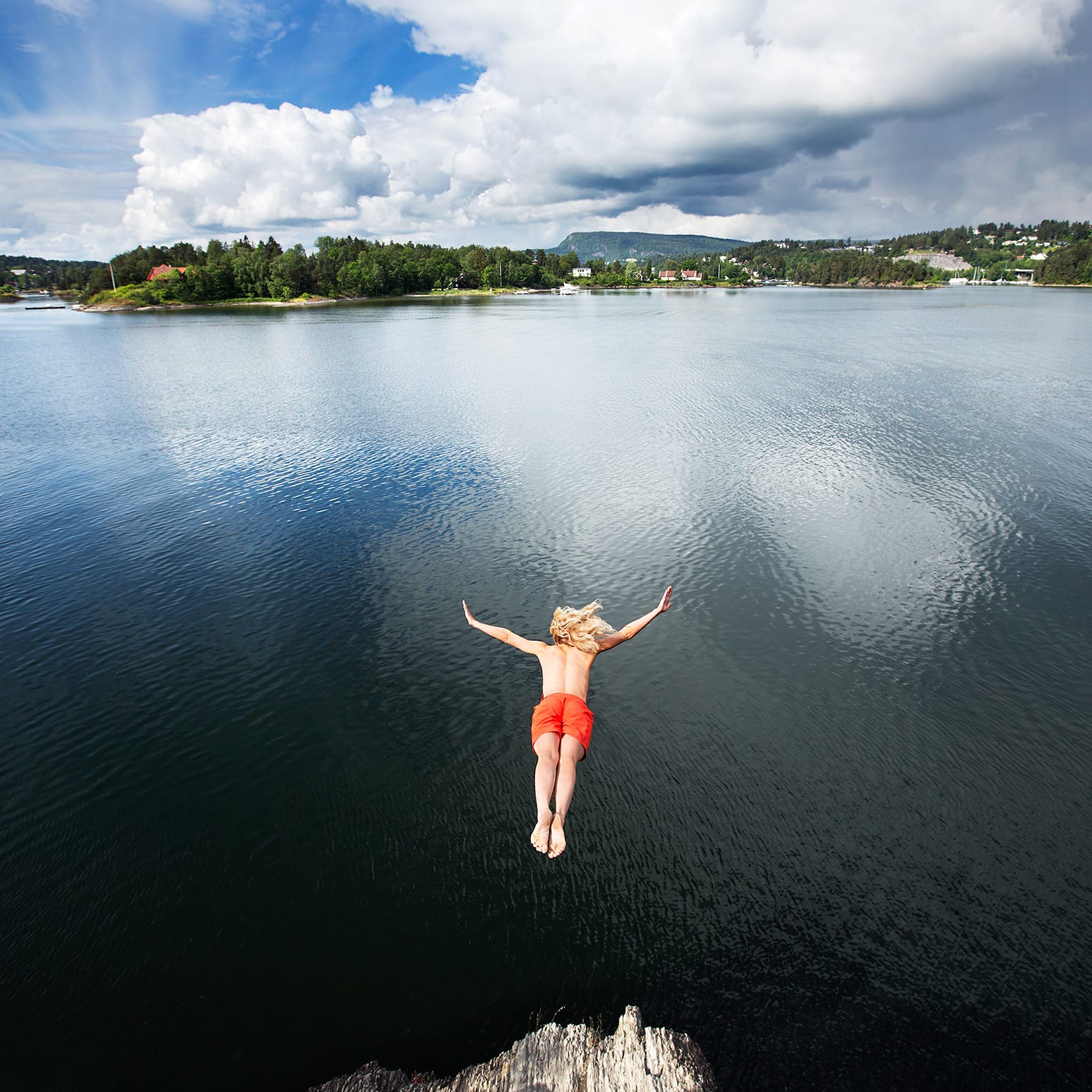 A person wearing red Norrøna shorts jumps into a body of water. The water is calm and the sky is cloudy.