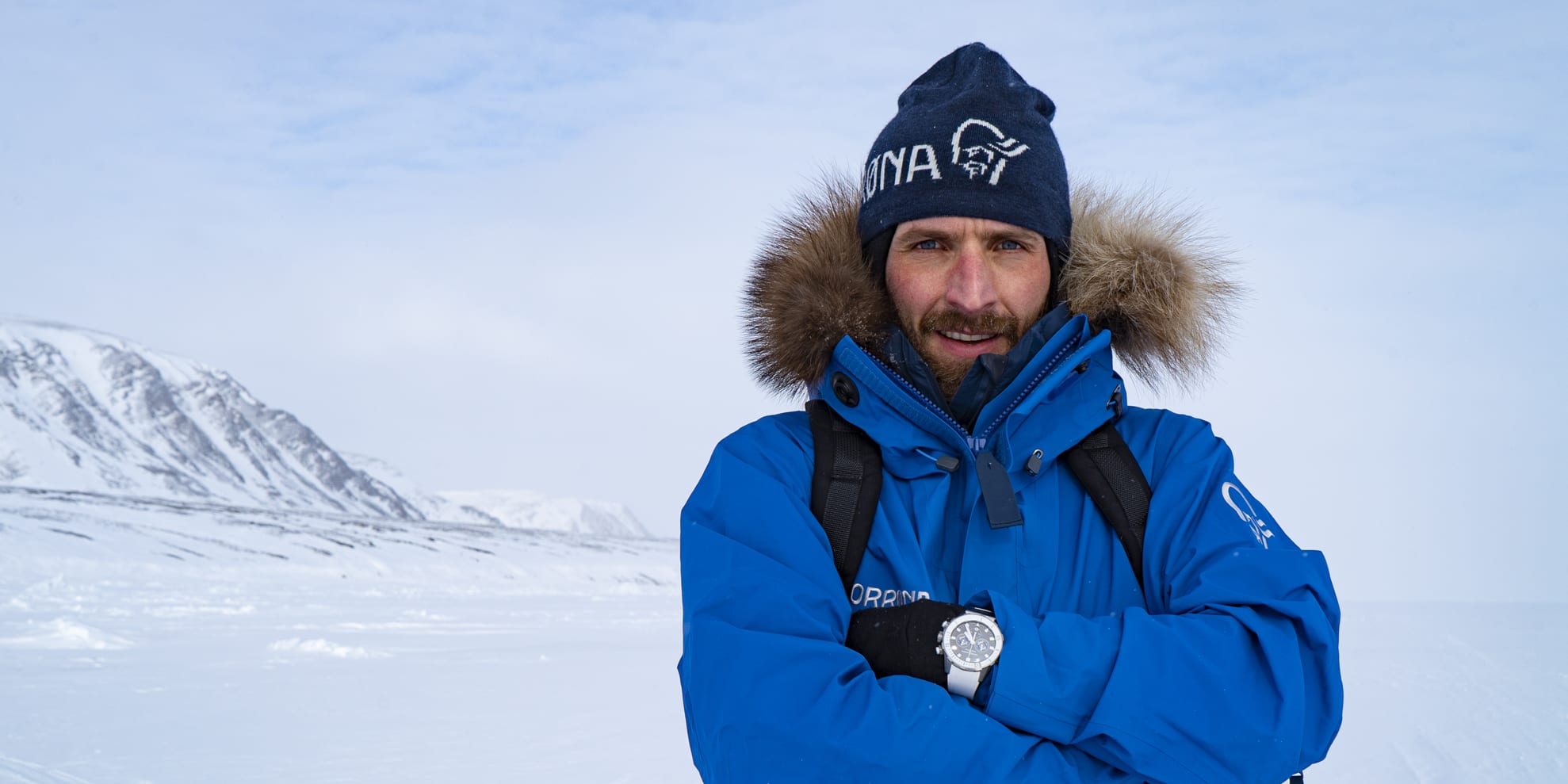 A man wearing a Norrøna jacket and hat stands in a snowy landscape. He is smiling and has his arms crossed. A red bag sits in the snow behind him.