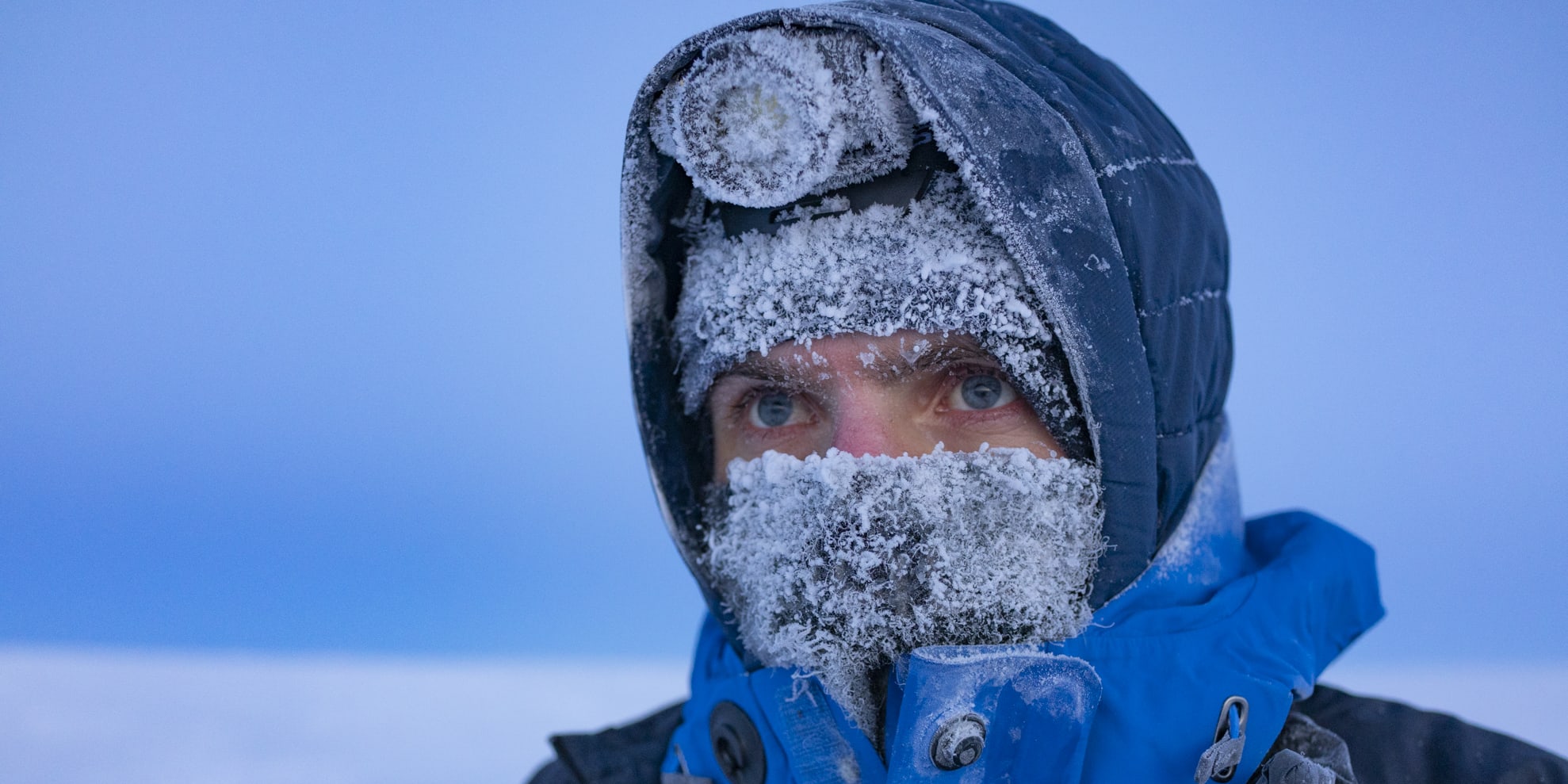 A person wearing a blue Norrøna jacket, hood, and a black Norrøna vest, is covered in frost. Their face is obscured by the frost and their eyes are visible.