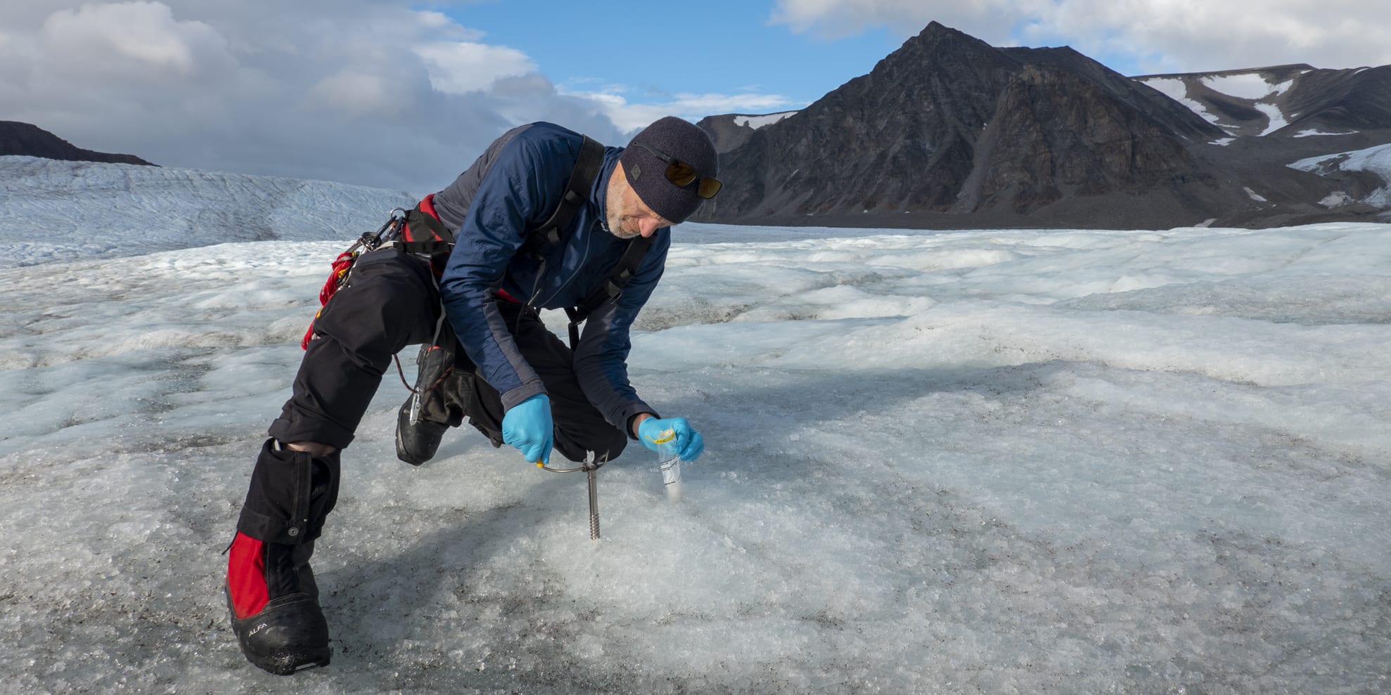 A person wearing Norrøna clothing kneels on a glacier, holding a small sample vial and using a metal probe to take a sample.  In the distance, a mountain range rises up against a clear blue sky. 
