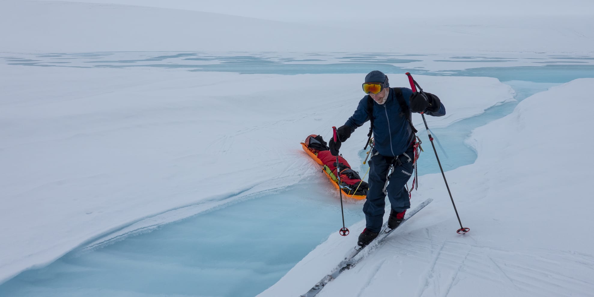 A person wearing ski gear, including a Norrøna jacket and pants, skis across a snowy landscape. They are pulling a bright orange sled with a red sleeping bag on it. The sled is sitting in a patch of blue ice.  