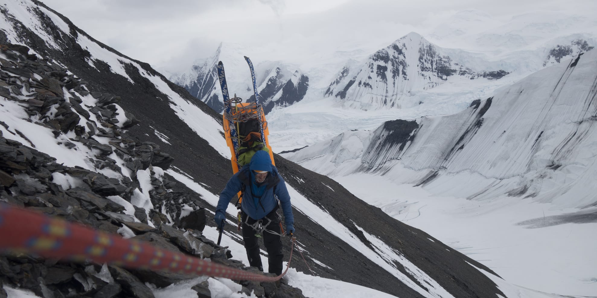 A person wearing Norrøna clothing,  climbing a snowy mountainside. They are carrying a bright orange sled with skis. The person is roped to the mountain and is wearing a helmet. The image shows a steep snowy mountainside with a glacier in the background.  