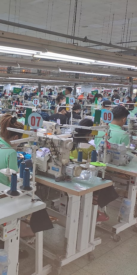 A wide shot of a sewing factory with many workers, all wearing face masks, operating sewing machines.  The workers are all wearing Norrøna clothes. 

