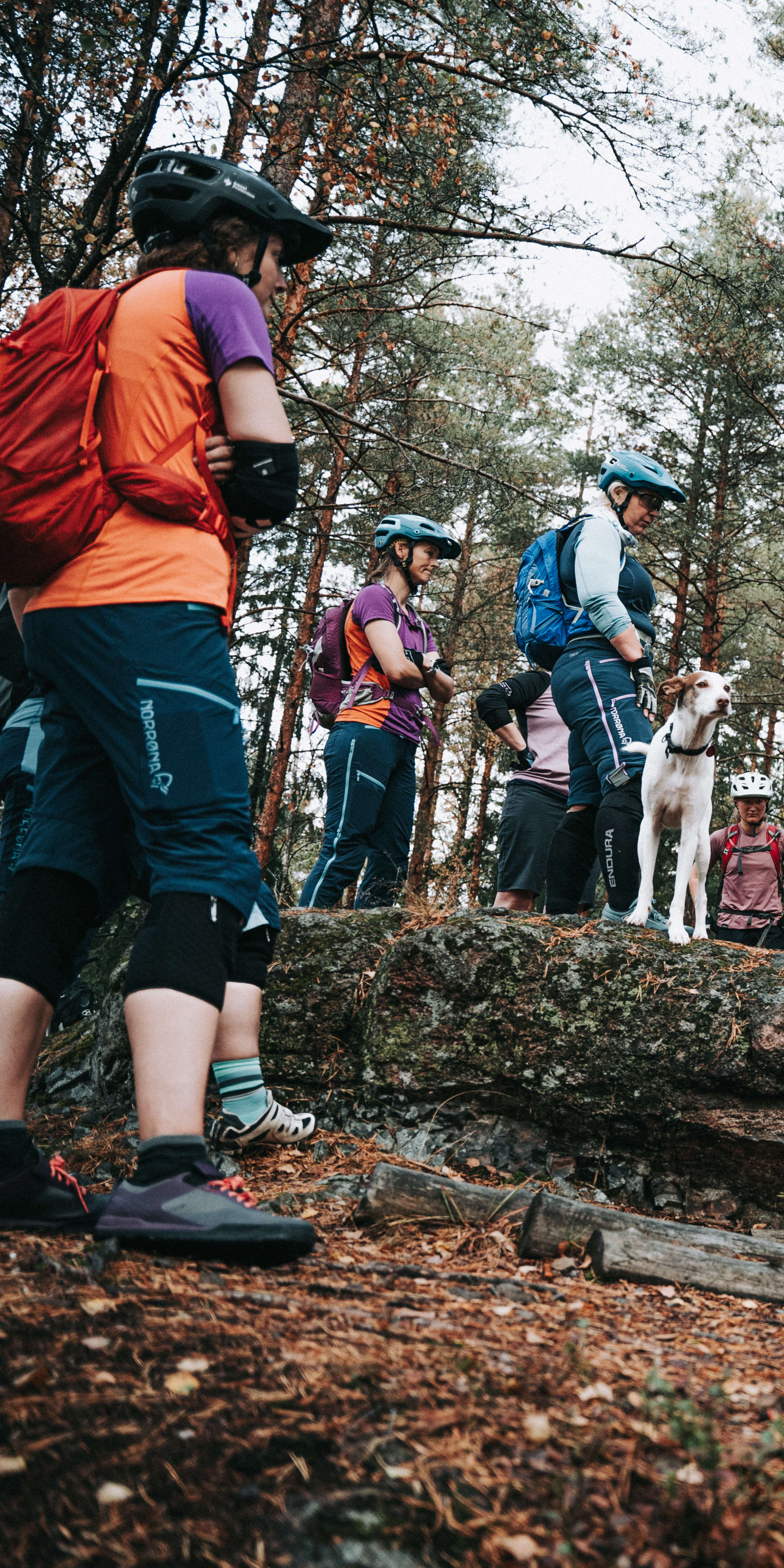 Group of people wearing helmets and Norrøna clothing in a forest setting with a mountain bike.  
