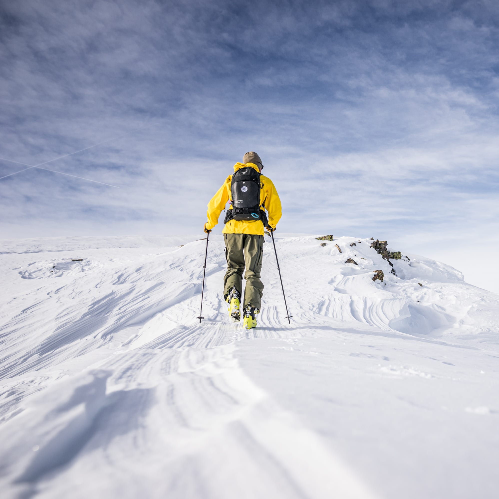 A person in a Norrøna yellow jacket and black backpack ski-touring in the snowy mountains, seen from behind. Blue sky above.