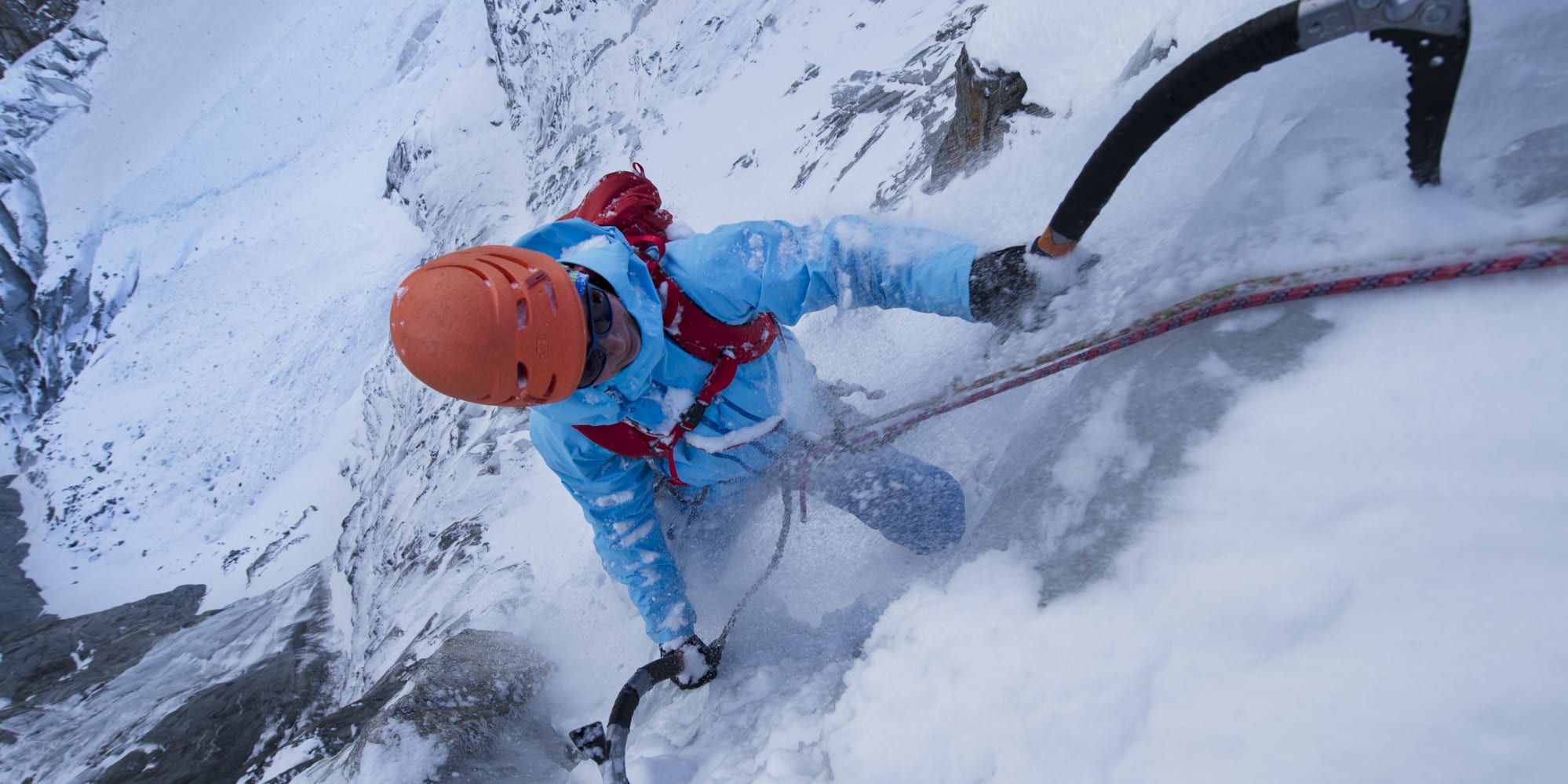 A climber, wearing Norrøna clothing and an orange helmet, ascends a steep, snowy mountainside using ice axes and a red and white rope.
