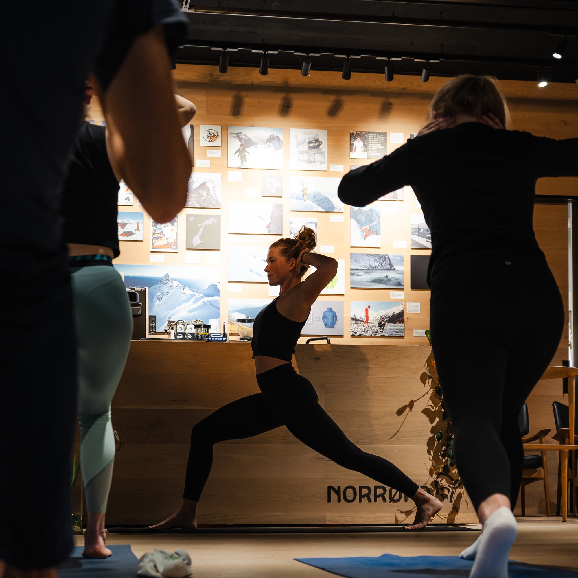 A yoga class in progress. A woman in Norrøna workout clothes strikes a warrior pose on a blue yoga mat.