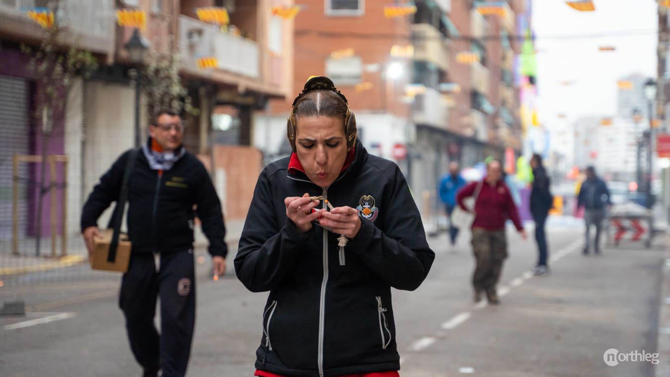 A person in a black jacket holding a small firecracker on a street during Valencia's Las Fallas festival.