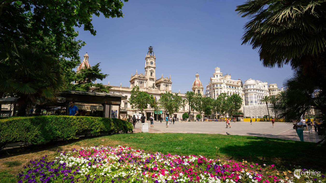 Panoramic view of Valencia's Plaza del Ayuntamiento with historic buildings, colorful flower beds, and palm trees under a clear blue sky.