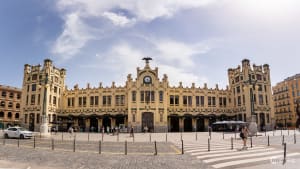 Exterior view of Valencia's North Station, a grand yellow-toned modernist building with decorative towers and a central clock, surrounded by an open plaza.