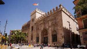 Exterior view of La Lonja de la Seda, a grand Gothic-style stone building with crenellated walls and ornate windows, located in a bustling Valencia street with tourists and yellow barriers.
