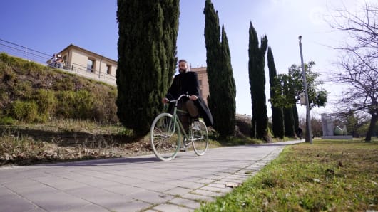 A cyclist riding a light green bicycle along a paved path lined with tall cypress trees and urban buildings in Valencia.