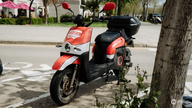 Red and black Acciona electric scooter parked on a city street with trees and urban background