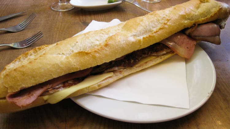 A long baguette sandwich with ham, cheese, and other fillings, served on a white plate with forks nearby.