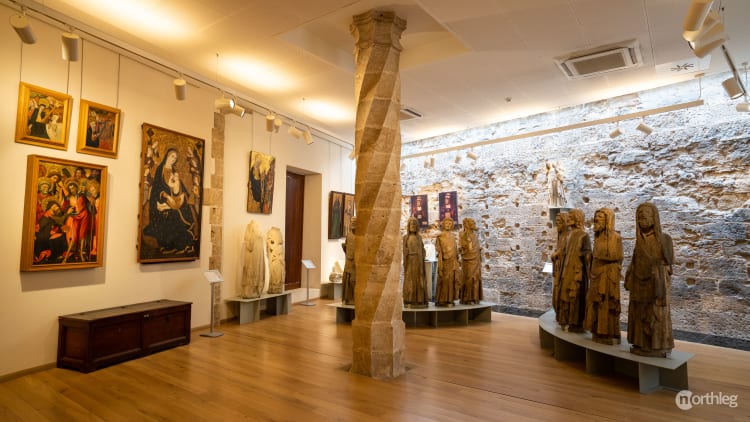 Interior museum room with medieval religious paintings, stone column, and wooden statues displayed on curved platforms against a stone wall.