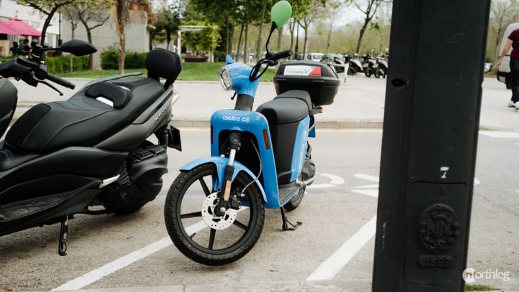 Blue Cooltra electric scooter parked in a designated parking area with other scooters and trees in the background.