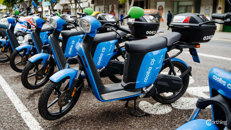 Blue Cooltra electric scooters parked in designated street parking spaces, featuring numbered vehicles with green helmets.