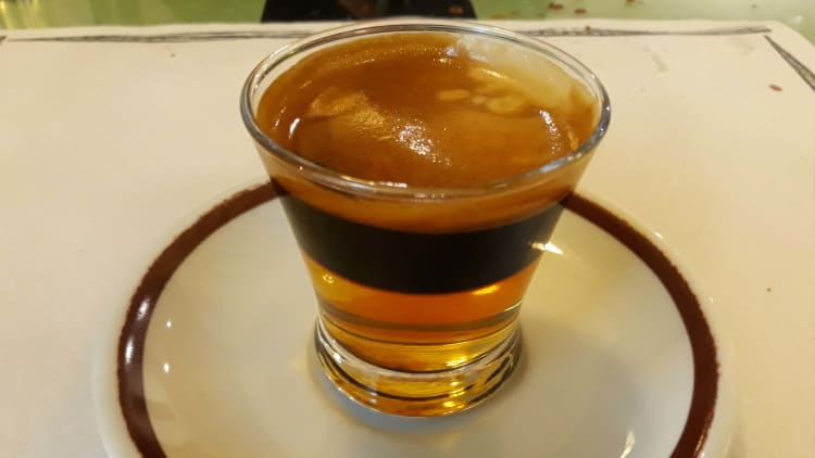 Layered golden and dark brown Cremaet cocktail in a clear glass, served on a white plate with a brown rim.