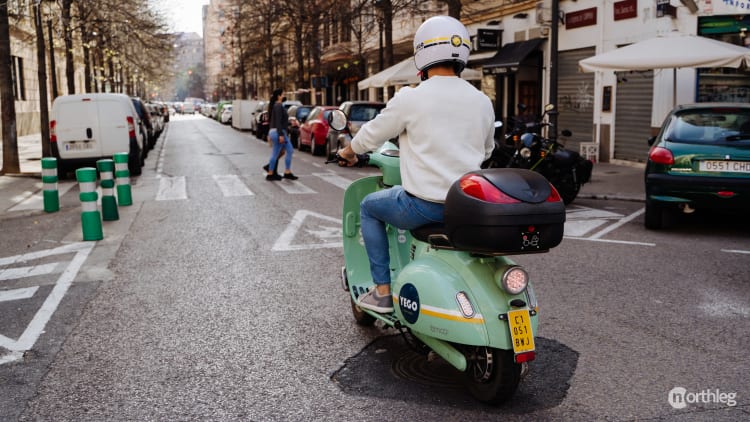 Green electric moped parked on a street in Valencia, with a rider wearing a white helmet