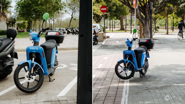 Two blue electric scooters, one parked correctly in a marked parking zone and the other blocking a pedestrian walkway.