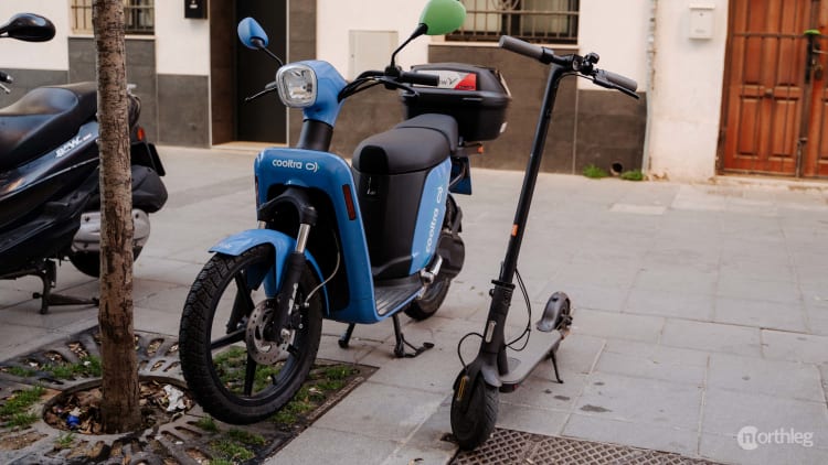 Blue seated electric scooter and gray stand-up electric scooter parked on a street in Valencia, Spain.