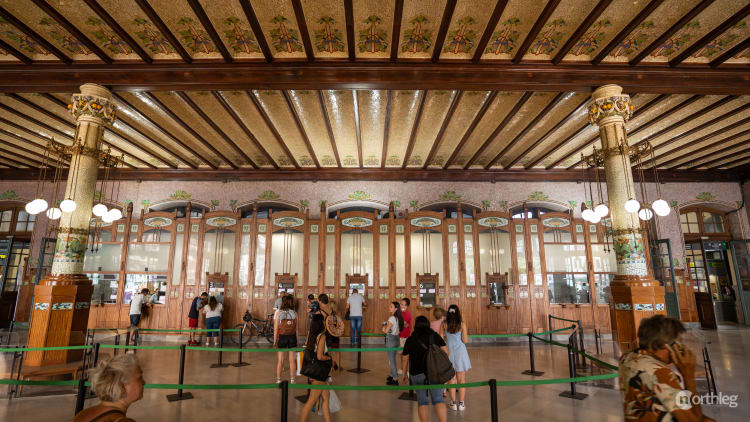 Ornate wooden ticket office with decorative ceiling and columns, featuring multiple service windows and people queuing in a grand historic train station interior.