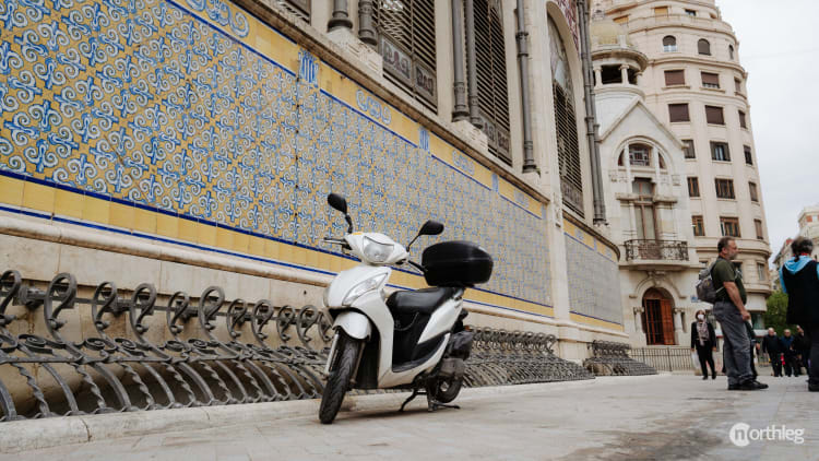 White electric scooter parked in front of a decorative tiled wall near the Valencia Central Market, with ornate architectural details visible.