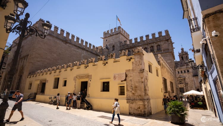 Exterior view of a yellow and stone Gothic-style building with crenellated walls, surrounded by historic streets and tourists on a sunny day.
