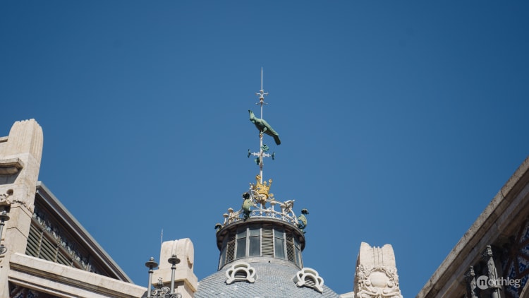 Bronze parrot weathervane on an ornate market building roof against a bright blue sky